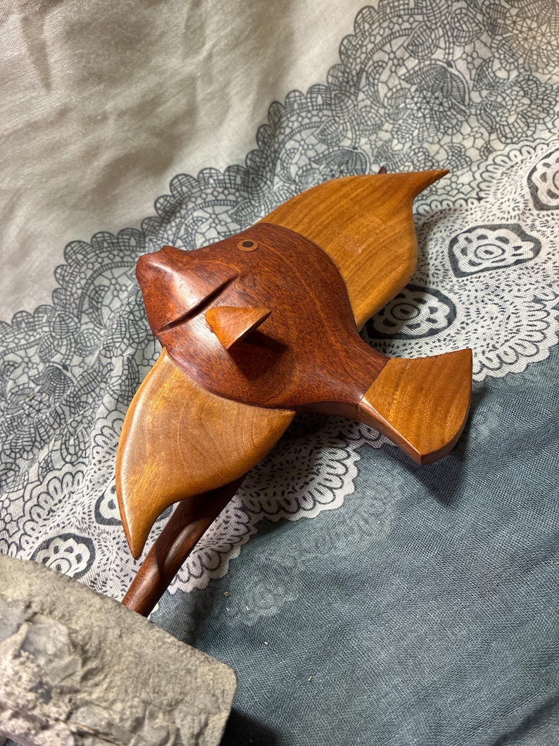 May include: A wooden fish sculpture with a detailed design. The fish is crafted from two shades of wood, with a darker brown head and a lighter brown body and fins. The sculpture has a whimsical, handcrafted appearance.