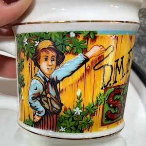 May include: White ceramic mug with a gold rim and a detailed illustration. The design features a young boy in a hat, writing on a wooden fence. The fence is yellow, and the boy is surrounded by green foliage. The mug has the letters "D.M.E.S."