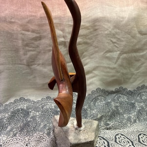 May include: A wooden sculpture featuring two abstract forms. One is a curved, dark brown shape, and the other is a lighter brown, wing-like form. The sculpture is mounted on a small, gray stone base. The background is a soft, neutral color.