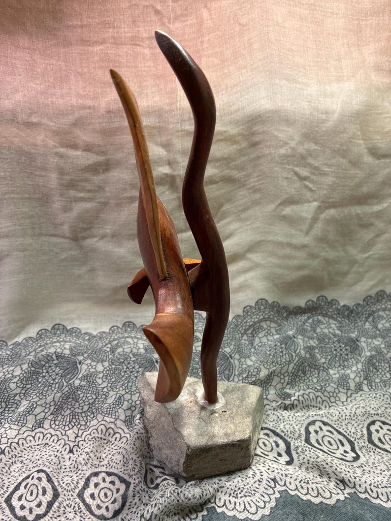 May include: A wooden sculpture featuring two abstract forms. One is a curved, dark brown shape, and the other is a lighter brown, wing-like form. The sculpture is mounted on a small, gray stone base. The background is a soft, neutral color.