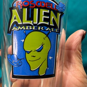 May include: Clear glass with a colorful graphic for Roswell Alien Amber Ale. The design features a green alien head with blue eyes, a blue UFO, and the words "Roswell Alien Amber Ale" in red, green, and white.