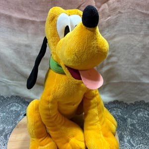 Op de afbeelding: Een pluche speelgoed van Pluto, een gele hond met zwarte oren en neus, witte ogen en een roze tong. Hij draagt een groene halsband. Het knuffeldier zit op een houten oppervlak.