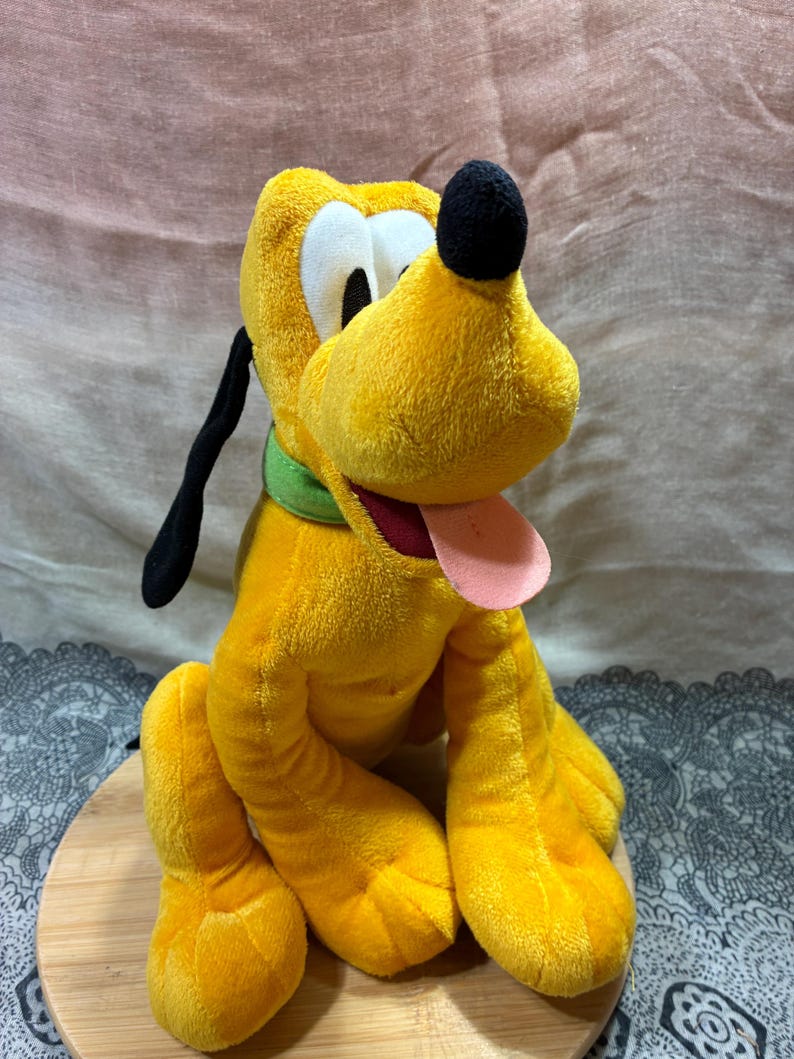 Op de afbeelding: Een pluche speelgoed van Pluto, een gele hond met zwarte oren en neus, witte ogen en een roze tong. Hij draagt een groene halsband. Het knuffeldier zit op een houten oppervlak.