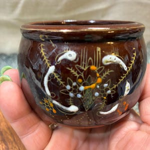 May include: A small, round, brown ceramic pot with a glossy finish. The pot features a floral design in white, orange, and blue. The rim is trimmed with gold. This decorative pot is ideal for small plants or as a decorative accent.