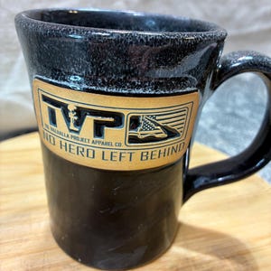May include: Black ceramic mug with a handle. The mug features a gold-colored rectangular emblem with the text "TVP" and "NO HERO LEFT BEHIND". The emblem also includes an American flag graphic. The mug is sitting on a wooden surface.
