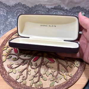May include: An open, brown Anne Klein glasses case with a beige interior. The brand name is printed in black. The case is held over a wooden, circular decorative piece with a tree branch design.