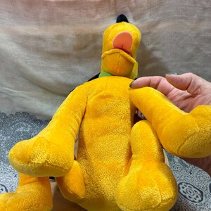 Op de afbeelding: Een pluche speelgoed van Pluto, een gele hond met zwarte oren, een groene kraag en een roze tong. Het knuffeldier zit op een houten oppervlak.
