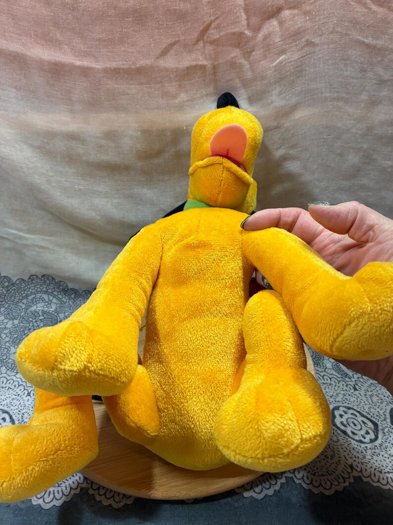Op de afbeelding: Een pluche speelgoed van Pluto, een gele hond met zwarte oren, een groene kraag en een roze tong. Het knuffeldier zit op een houten oppervlak.