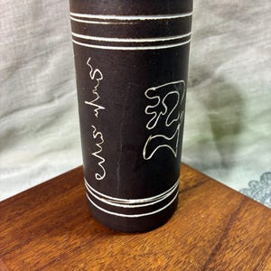 May include: A dark brown ceramic tumbler with white etched designs and text. The tumbler has two white bands near the top and bottom. The tumbler has a white etched design and text on the side.