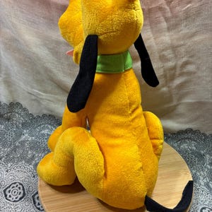 Op de afbeelding: Een pluche speelgoed van Pluto, een gele hond met zwarte oren en staart, een groene kraag en een roze tong. Het knuffeldier zit op een houten oppervlak.