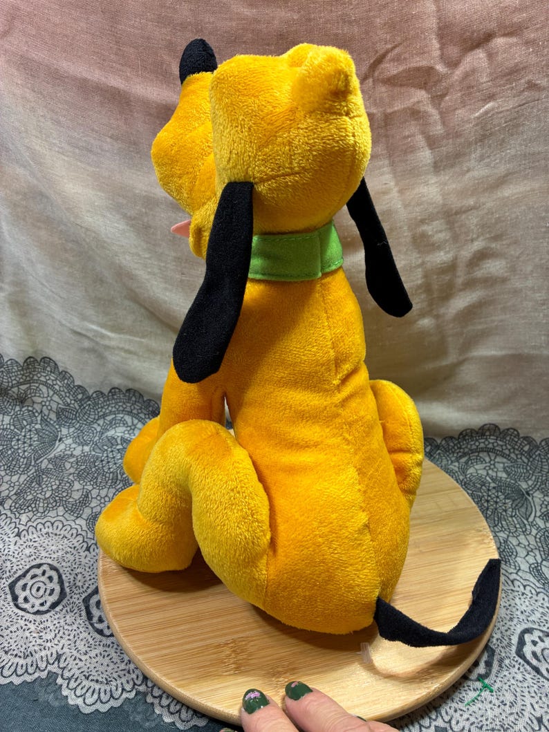 Op de afbeelding: Een pluche speelgoed van Pluto, een gele hond met zwarte oren en staart, een groene kraag en een roze tong. Het knuffeldier zit op een houten oppervlak.