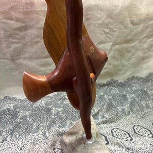 May include: A wooden sculpture of a fish with a dark brown body and fins, and a lighter brown wing. The fish is mounted on a gray stone base. The sculpture has a smooth finish and is likely handmade.