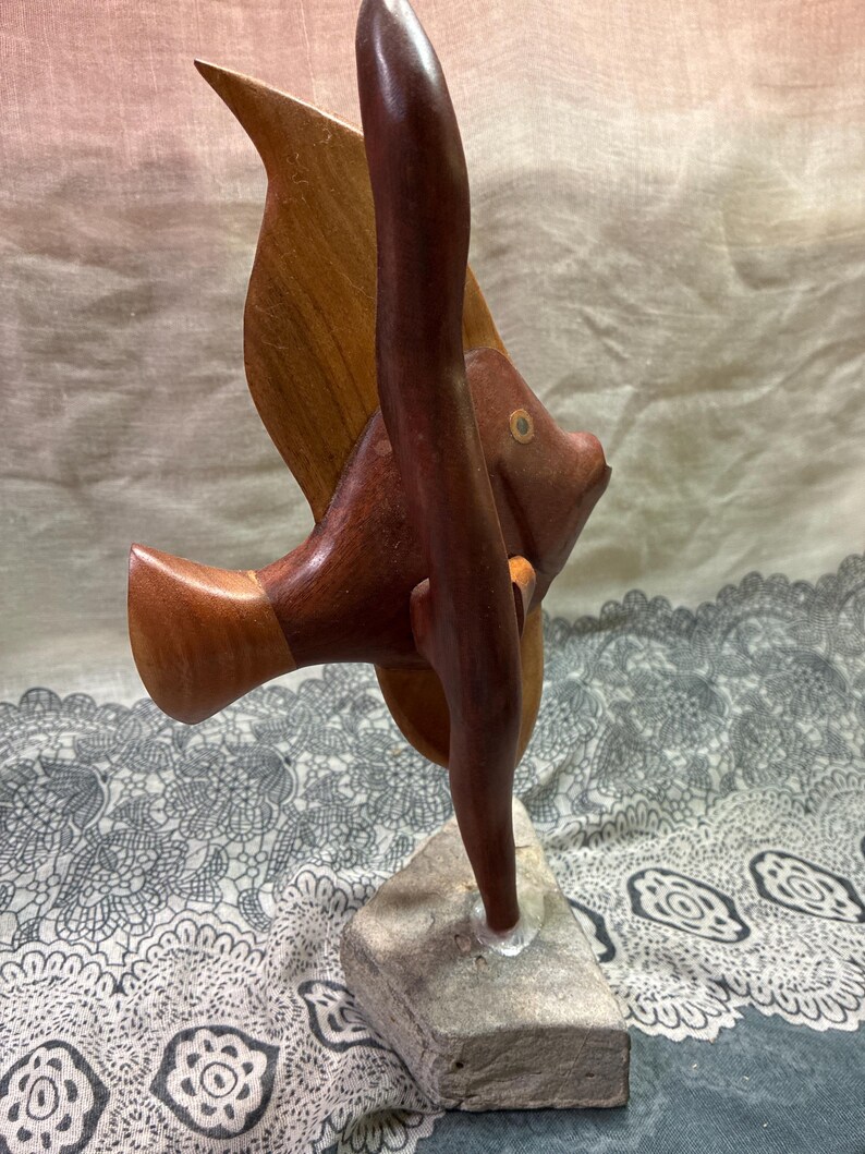 May include: A wooden sculpture of a fish with a dark brown body and fins, and a lighter brown wing. The fish is mounted on a gray stone base. The sculpture has a smooth finish and is likely handmade.