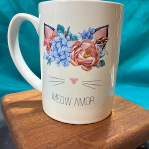 May include: White ceramic mug with a cat face design. The design includes pink cat ears, a floral arrangement of blue and red flowers, and the text "MEOW AMOR" below the cat face. The mug has a curved handle.