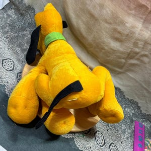Op de afbeelding: Een pluche geel hondenspeelgoed met zwarte oren en een groene kraag. De hond zit op een houten voet. Het speelgoed is een klassiek stripfiguur.