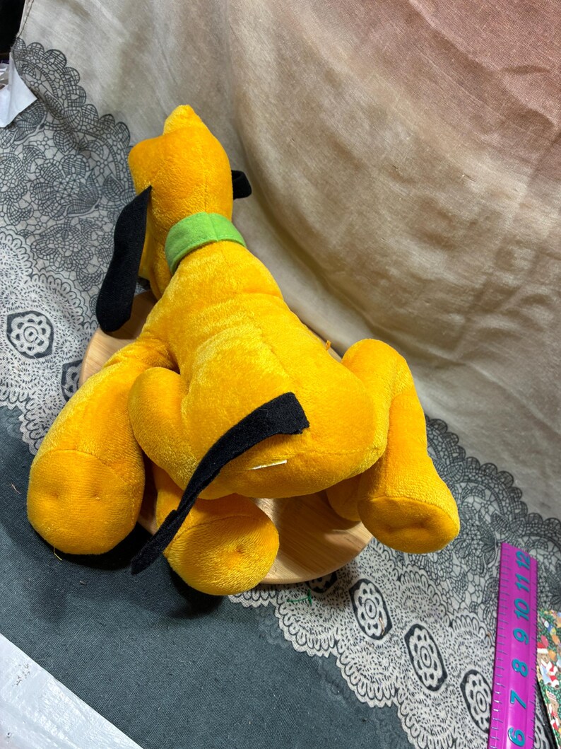Op de afbeelding: Een pluche geel hondenspeelgoed met zwarte oren en een groene kraag. De hond zit op een houten voet. Het speelgoed is een klassiek stripfiguur.