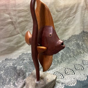 May include: A handcrafted wooden fish sculpture with a dark brown body and lighter brown fins. The fish is mounted on a gray stone base. The sculpture has a unique, artistic design, showcasing the natural wood grain.