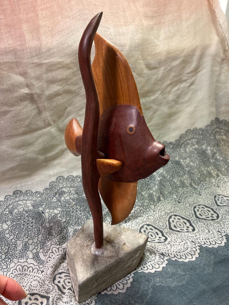 May include: A handcrafted wooden fish sculpture with a dark brown body and lighter brown fins. The fish is mounted on a gray stone base. The sculpture has a unique, artistic design, showcasing the natural wood grain.