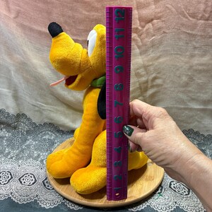 Op de afbeelding: Een pluche Pluto-hondenspeelgoed, voornamelijk geel met zwarte en witte accenten, wordt getoond op een houten basis. Het speelgoed is ongeveer 28 cm hoog, gemeten met een paarse liniaal. De hond heeft een groene kraag en een roze tong.