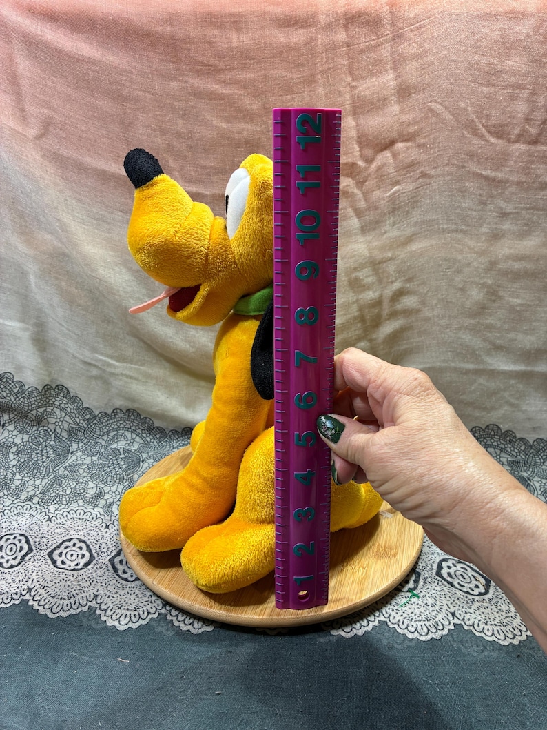 Op de afbeelding: Een pluche Pluto-hondenspeelgoed, voornamelijk geel met zwarte en witte accenten, wordt getoond op een houten basis. Het speelgoed is ongeveer 28 cm hoog, gemeten met een paarse liniaal. De hond heeft een groene kraag en een roze tong.
