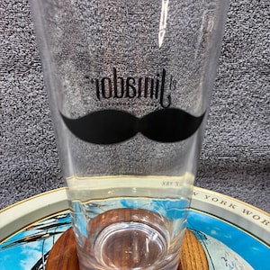 May include: A clear pint glass with a black mustache graphic and the word "robsmit" in a stylized font. The glass is on a wooden coaster, with a vintage tray in the background.