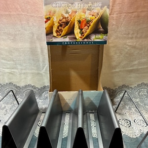 May include: A metal taco rack with four slots, displayed in front of a Chicago Metallic box. The box features images of tacos and the text "Chicago Metallic The Baking Experts PROFESSIONAL". The rack is made of silver-colored metal.