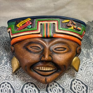 May include: A brown ceramic mask with a smiling face and gold-colored earrings. The mask has a colorful headdress with green, red, orange, and yellow accents. The mask is likely a decorative item or wall hanging.