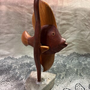 May include: A wooden sculpture of a fish with a dark brown body and light brown fins. The fish is mounted on a small, gray stone base. The sculpture has a smooth finish and is likely handmade.
