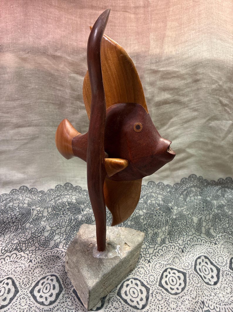 May include: A wooden sculpture of a fish with a dark brown body and light brown fins. The fish is mounted on a small, gray stone base. The sculpture has a smooth finish and is likely handmade.
