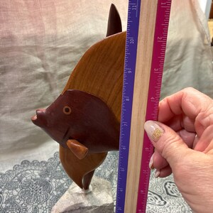 May include: A wooden fish sculpture, approximately 10 inches tall, with a dark brown head and a lighter brown body. The sculpture is mounted on a gray stone base. The fish has a detailed design and is a decorative item.