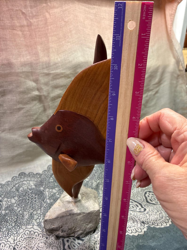 May include: A wooden fish sculpture, approximately 10 inches tall, with a dark brown head and a lighter brown body. The sculpture is mounted on a gray stone base. The fish has a detailed design and is a decorative item.