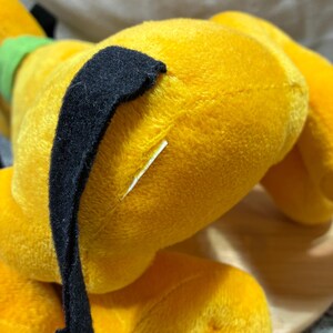 Op de afbeelding: Een gele Pluto-knuffel, de Disney-tekenfilmhond, in liggende positie. Het heeft een zwart oor en een groene kraag. Het speelgoed is gemaakt van zacht, pluizig materiaal.