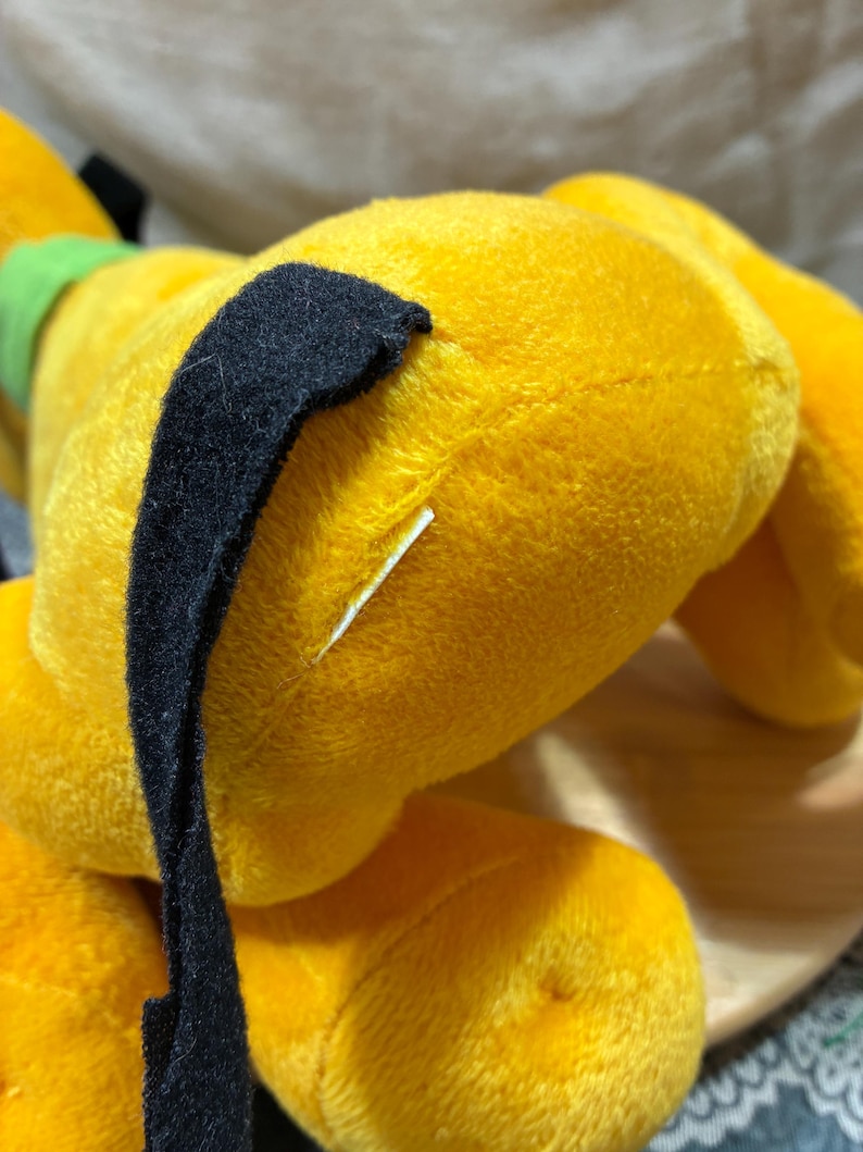 Op de afbeelding: Een gele Pluto-knuffel, de Disney-tekenfilmhond, in liggende positie. Het heeft een zwart oor en een groene kraag. Het speelgoed is gemaakt van zacht, pluizig materiaal.