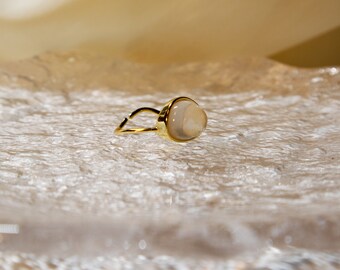 May include: A gold ring with a white oval-shaped gemstone. The ring is on a white textured surface.