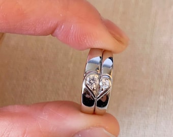 Könnte beinhalten: Zwei silberne Eheringe mit einem Herzdesign und kleinen Diamanten. Die Ringe werden von einem Finger zusammengehalten.