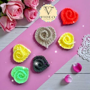 May include: A collection of handmade crochet heart-shaped appliques in various colors, including red, yellow, green, gray, and black. The appliques are arranged on a pink surface with rose petals and a logo that says "VIDEO TUTORIAL".