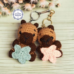 May include: Two crocheted capybara keychains with dark brown snouts and light brown bodies. One capybara holds a light blue crocheted star, and the other holds a light pink crocheted star. Both feature black plastic eyes and metal key rings.