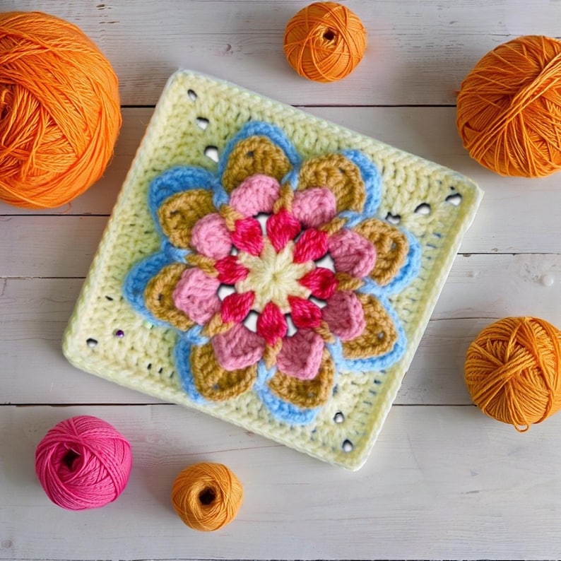 May include: A square, crocheted flower motif in a variety of colors, including pink, blue, and gold, sits on a light yellow background. Several balls of orange and pink yarn are scattered around the motif. The handmade item is likely part of a larger craft project.