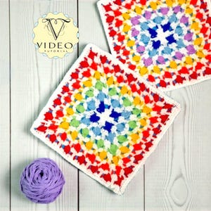 May include: Two colorful, square crochet pieces with a rainbow pattern and white borders. A ball of purple yarn sits near one of the squares. The image also includes a logo with the text "VIDEO TUTORIAL".