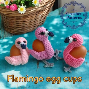 May include: Three pink crocheted flamingo egg cups holding brown eggs. The egg cups are on a blue and white patterned surface. The text "Flamingo egg cups" is at the bottom of the image.