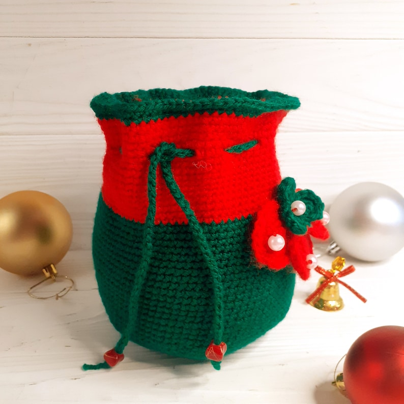 K&ouml;nnte beinhalten: Eine gr&uuml;ne und rote geh&auml;kelte Kordeltasche mit einer wei&szlig;en Perlenverzierung. Die Tasche liegt auf einer wei&szlig;en Holzoberfl&auml;che mit Weihnachtsornamenten.