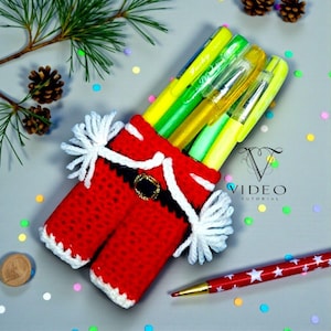 May include: A handmade red and white crochet pen holder shaped like Santa's pants, filled with colorful pens and highlighters. The holder has a black belt with a gold buckle and white trim. A red pen with white stars is also visible.