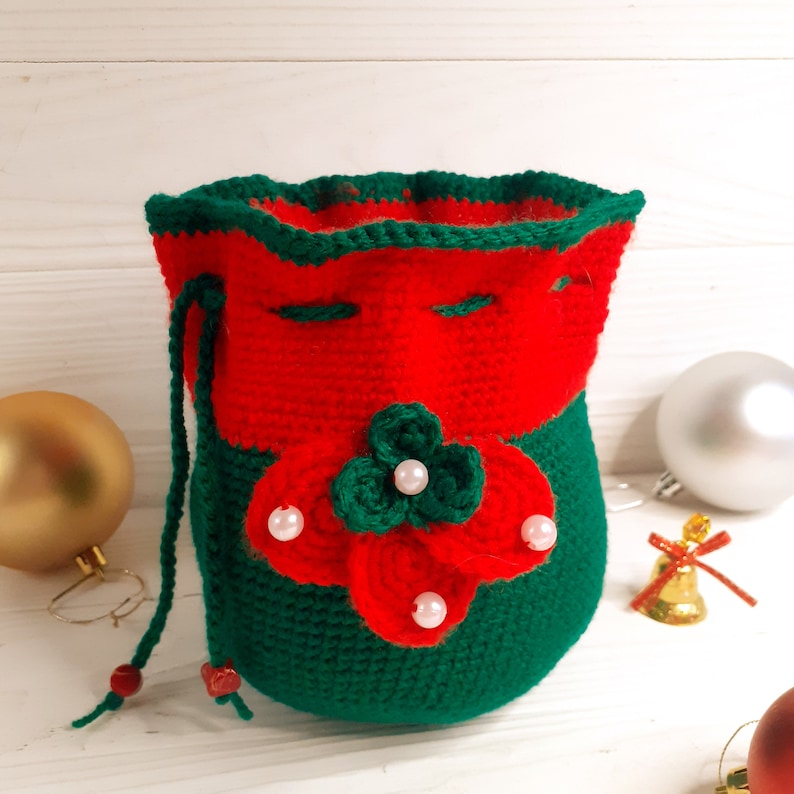 K&ouml;nnte beinhalten: Eine gr&uuml;ne und rote geh&auml;kelte Kordeltasche mit Blumendesign und wei&szlig;en Perlenakzenten. Die Tasche ist perfekt f&uuml;r kleine Geschenke oder Kleinigkeiten.