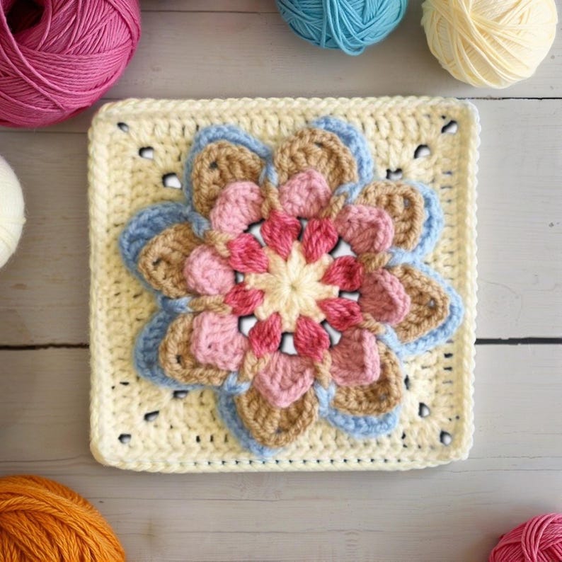 May include: A square, crocheted flower motif in shades of pink, tan, and blue, centered on a cream-colored square. The flower has multiple layers of petals, with a light yellow center. Yarn balls in pink, blue, and yellow are in the background.