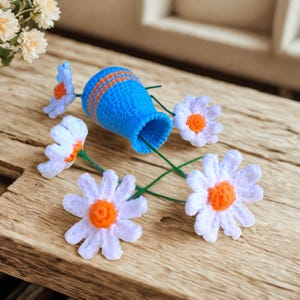 Könnte beinhalten: Eine handgefertigte Häkelvase in leuchtendem Blau mit orangefarbenen Streifen, umgeben von weißen und orangefarbenen Häkelgänseblümchen mit grünen Stielen. Die Blumen sind auf einer Holzoberfläche angeordnet und schaffen eine charmante, skurrile Präsentation.