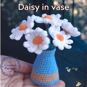 May include: A crocheted bouquet of white daisies with orange centers in a blue vase with orange stripes. The vase is crocheted and the flowers are arranged in a vase.
