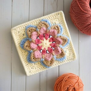May include: A square, crocheted flower motif in cream, with a central flower design in pink, light brown, and blue. Two balls of orange yarn are in the background. The crochet work is detailed and the colors are soft.
