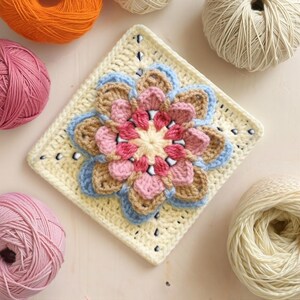 May include: A colorful crochet flower square in shades of pink, blue, tan, and cream. The square is surrounded by balls of yarn in various colors, including pink, orange, and cream. This handmade item is perfect for crafting projects.