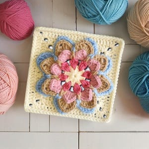 May include: A square, crocheted flower motif in shades of pink, beige, and blue, centered on a pale yellow square. Several balls of yarn in coordinating colors surround the motif, suggesting a craft project. The handmade item is likely part of a larger textile creation.