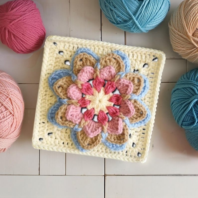 May include: A square, crocheted flower motif in shades of pink, beige, and blue, centered on a pale yellow square. Several balls of yarn in coordinating colors surround the motif, suggesting a craft project. The handmade item is likely part of a larger textile creation.